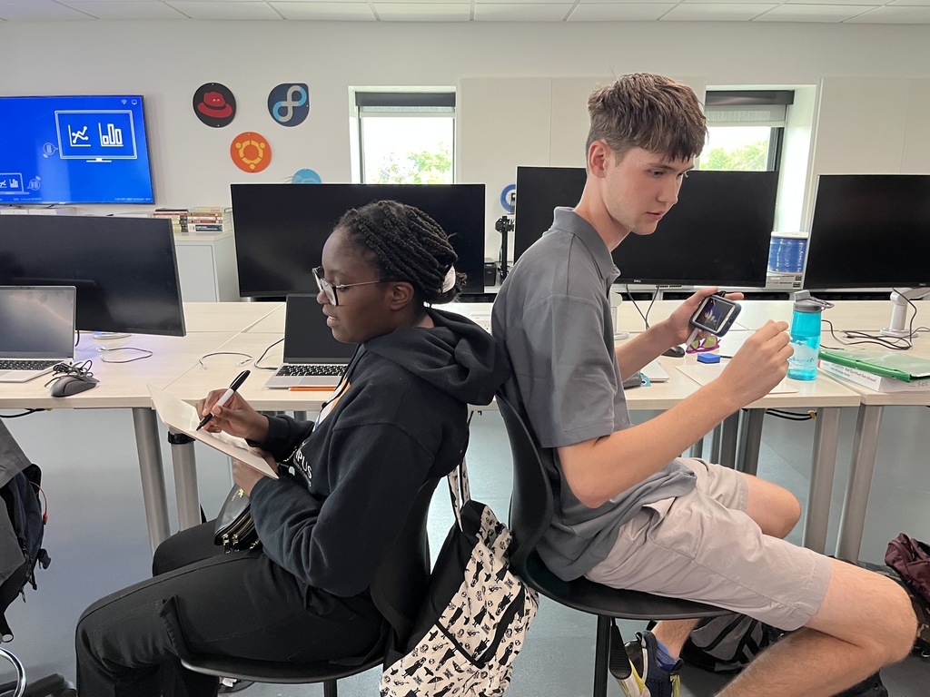 Two high school students sit back-to-back on chairs in a computer lab. The student on the left, a Black girl with glasses and braids, wears a black hoodie and writes in a notebook. The student on the right, a white boy in a gray polo and light shorts, holds a smartphone displaying a graph. They are surrounded by computer monitors, laptops, and classroom supplies. Tech-themed wall decals and a large screen displaying data icons are visible in the background.