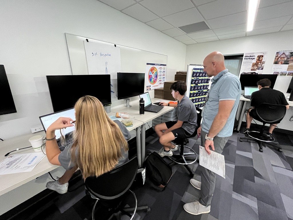 A classroom scene with students working individually on laptops at desks with large monitors. A teacher stands beside a male student, offering guidance. A female student with long blonde hair sits nearby, focusing on her work. Posters about design concepts and a perspective drawing hang on the walls, suggesting an architecture class.