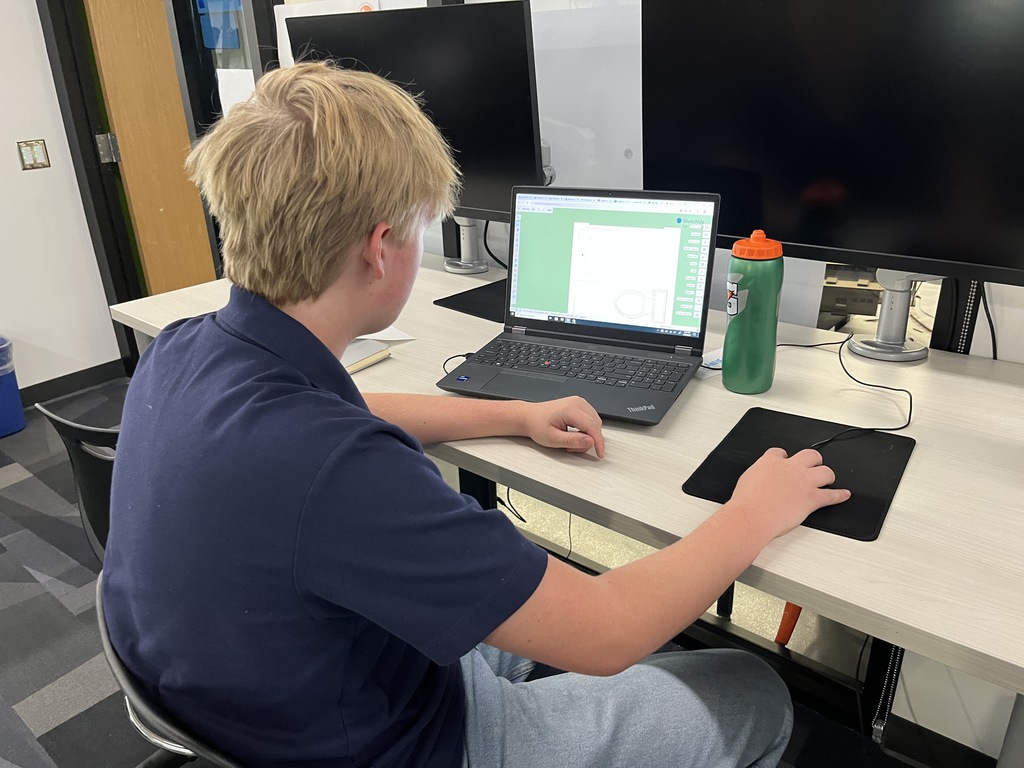 A blonde male student in a navy polo shirt works on a laptop at a classroom desk, using a mouse and looking at a CAD-style design on the screen. He is seated in front of multiple large monitors. A green water bottle with an orange cap sits beside the laptop. The setting is a tech-focused computer lab.
