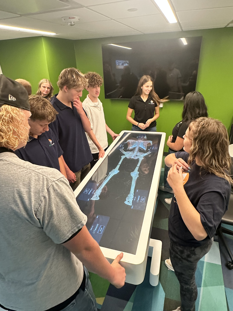A group of high school students gathers around an interactive digital anatomy table displaying a skeletal scan. The students, diverse in gender and appearance, are engaged and curious, with one student gesturing while others observe the screen. They wear casual and school-branded clothing. The room has a bright green wall, a large black screen mounted on it, and colorful geometric carpet tiles. Ceiling lights and reflections are visible above the display.
