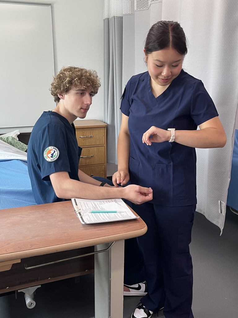 Two high school students in navy blue scrubs practice a healthcare scenario in a clinical classroom setting. One student, seated on a hospital bed, extends his wrist while the other stands beside him, checking the time on her smartwatch as if timing a pulse. A medical binder with a pen is on a nearby overbed table. The background includes privacy curtains, a wooden cabinet, and a neatly made hospital bed.