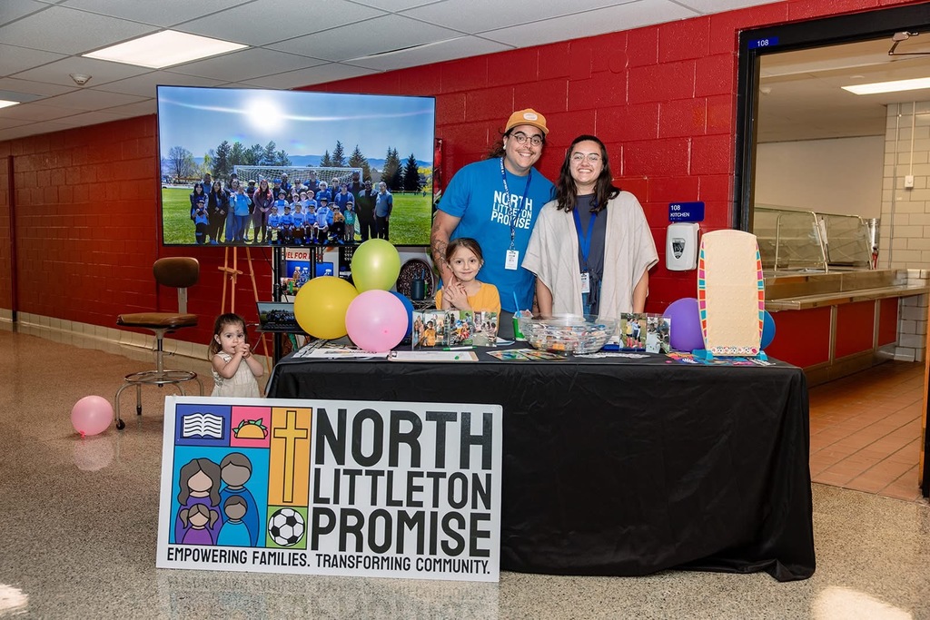 Two adults and two children at a North Littleton Promise table with balloons and display materials, promoting community programs.