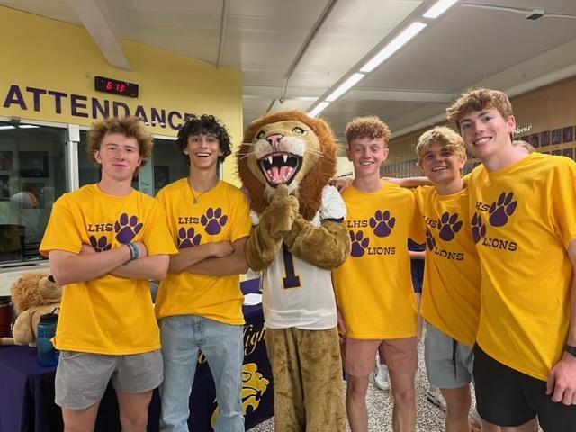 A group of six young men are standing indoors, smiling and posing for a photo with a person in a lion mascot costume. All five boys are wearing matching bright yellow "LHS Lions" t-shirts with purple paw prints, suggesting a school spirit or back-to-school event. They are standing in front of a purple table and a sign that reads "ATTENDANCE." The atmosphere is cheerful and energetic, indicating school pride and camaraderie.