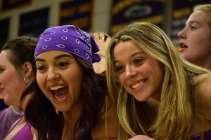Two enthusiastic young women are smiling and leaning forward, appearing to cheer or react excitedly at an event. One is wearing a purple bandana and a big open-mouthed smile, while the other has long blonde hair and is grinning brightly. They are surrounded by other students, and school banners hang in the background, suggesting a spirited school event or pep rally.