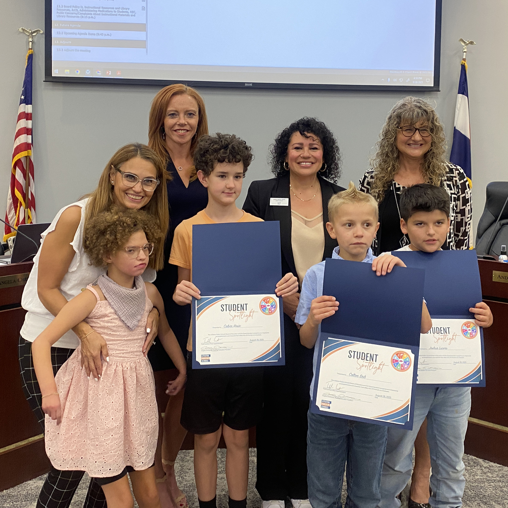 Four elementary students, 3 holding “Student Spotlight” certificates, pose with four smiling adults in a boardroom with flags and a screen.