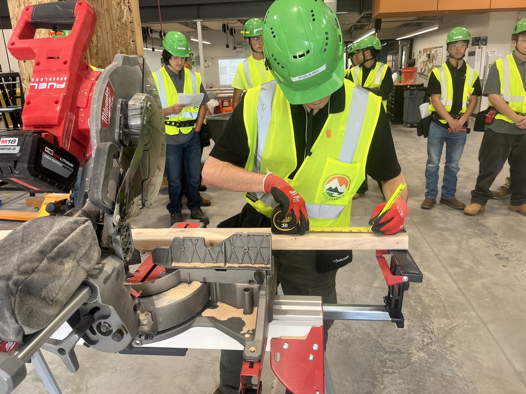 A student in a green hard hat, red gloves, and yellow safety vest measures a piece of wood with a tape measure at a miter saw station inside a construction classroom. The student is surrounded by peers also wearing safety gear, standing in the background observing. The vest features a circular patch with a mountain and sun logo. The setting is a modern workshop with concrete floors and visible equipment.