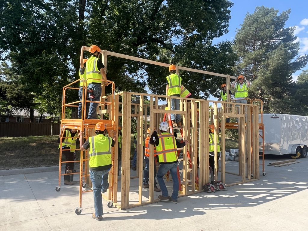 A group of students wearing orange hard hats and yellow safety vests construct a wooden frame structure outdoors under large trees. Some stand on orange scaffolding while others support the frame from the ground. In another scene, students work collaboratively on a concrete surface, measuring and aligning wooden boards. One student sits holding a blue triangle square while others crouch or stand nearby with tools. A partially built wooden structure is visible in the background.