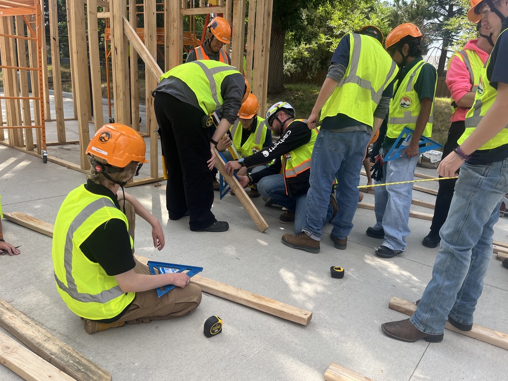 A group of students wearing orange hard hats and yellow safety vests construct a wooden frame structure outdoors under large trees. Some stand on orange scaffolding while others support the frame from the ground. In another scene, students work collaboratively on a concrete surface, measuring and aligning wooden boards. One student sits holding a blue triangle square while others crouch or stand nearby with tools. A partially built wooden structure is visible in the background.