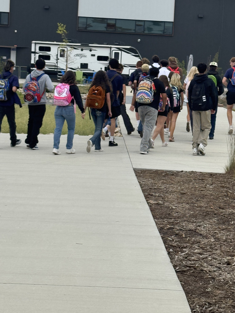 A group of diverse middle or high school students walking away on a wide sidewalk toward a modern school building. The students wear casual clothing and colorful backpacks with various designs, including a pink heart, cartoon characters, and a shark face. The group includes a mix of boys and girls with varied hair colors and styles. In the background, a white and black RV labeled “Imagine” is parked near the building, which has a dark exterior with large windows. The scene is outdoors with some mulch and grass along the path.