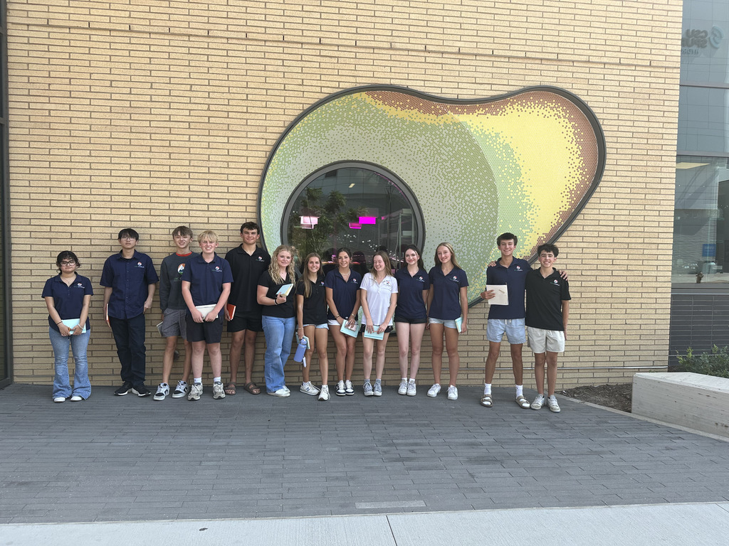 A group of thirteen high school students stands in a line outside a building with light tan brick walls. Behind them is a circular window framed by a large, avocado-shaped mosaic made of green, yellow, and brown tiles. Most students are wearing navy blue shirts with a small logo on the chest; a few wear black or white versions. They hold notebooks or papers and are smiling at the camera. The group is diverse in gender and appearance, and the setting appears to be an educational field trip, likely related to architecture or design based on the backdrop and materials in hand.