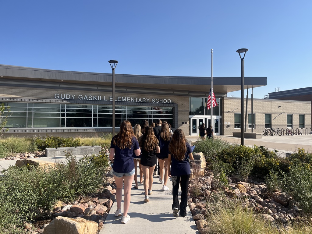 A group of students, mostly female and dressed in matching navy blue shirts, walks along a landscaped sidewalk toward the entrance of Gudy Gaskill Elementary School. The school name is prominently displayed above the front windows. The building is modern with tan brick walls and large glass windows. An American flag is flown at half-staff near the entrance. The surrounding landscape features rocks, native grasses, and low-maintenance plants. The weather is sunny with a clear blue sky.