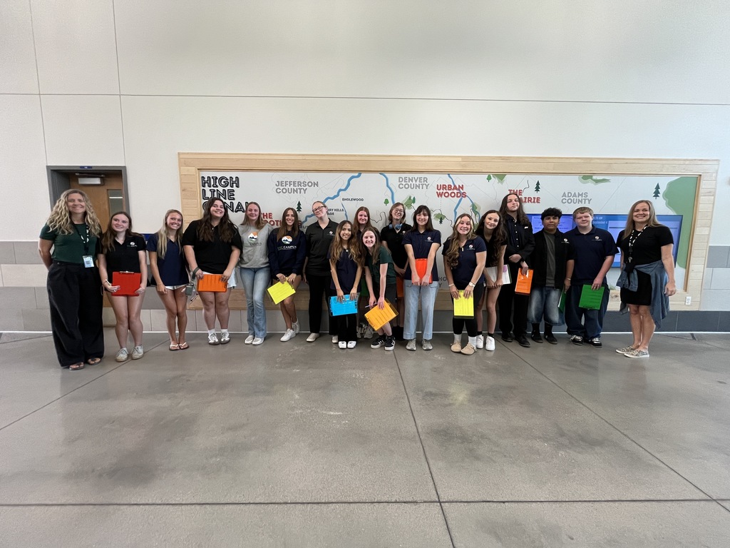A group of high school students and two adult staff members pose indoors in front of a large wall map labeled with local areas such as "High Line Canal," "Jefferson County," "Denver County," and "Urban Woods." The students, diverse in appearance, are smiling and holding colorful folders in red, orange, yellow, green, and blue. Most wear navy blue or black shirts with a small logo, and some are in jeans or casual attire. The setting is a school or educational facility with a clean, modern interior, gray flooring, and light-colored walls.