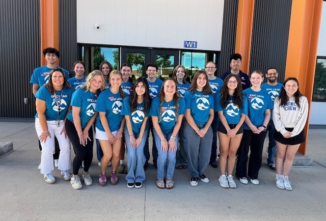 Group of 20 people, including diverse high school students and a few adults, posing and smiling in front of a modern school building with orange pillars and a sign labeled "W1." Most are wearing matching bright blue T-shirts with a white mountain logo and the words "EPIC LEAD." One person on the far right wears a gray "MINNESOTA CHEER" sweatshirt. The group stands on a concrete walkway with glass doors and windows behind them.