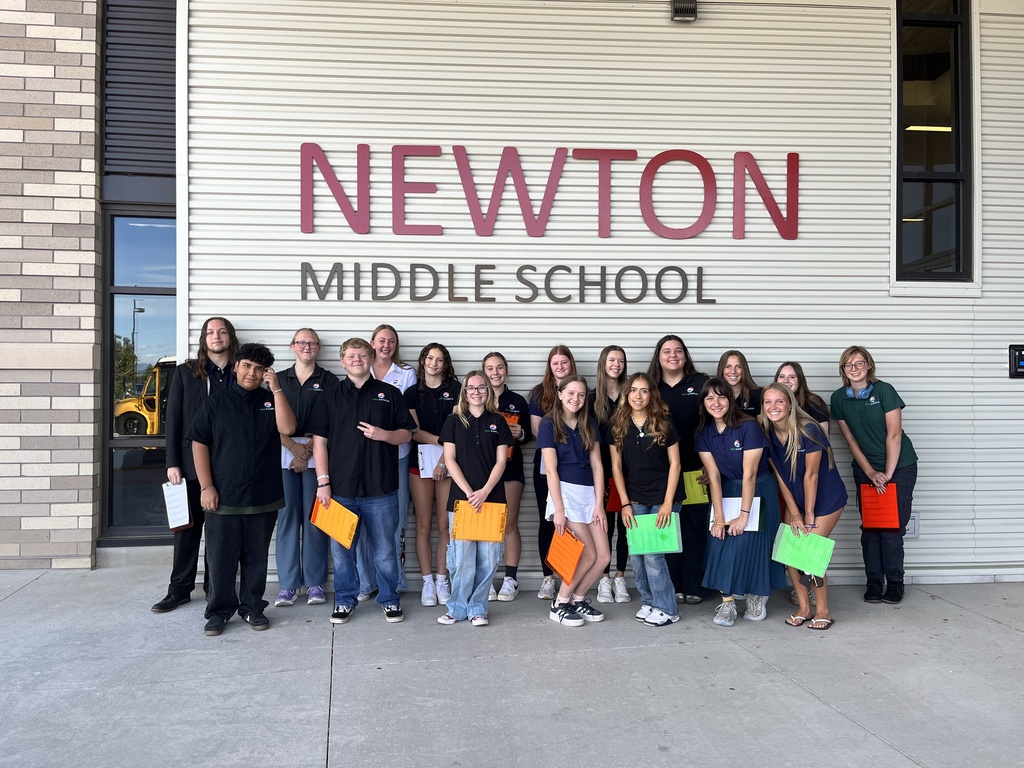 Group of 20 people, a mix of students and adults, standing in front of a wall labeled “NEWTON MIDDLE SCHOOL” in large red and gray letters. Most individuals are smiling and holding brightly colored folders or papers. The group is gathered on a concrete sidewalk next to a brick and metal-paneled school building. A school bus is partially visible through a window on the left.
