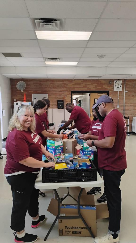 Volunteers in matching maroon shirts stand at a long table filled with various feminine hygiene products like pads and tampons. They are sorting and packing the items into boxes. One volunteer smiles at the camera while others work in the background. Cardboard boxes and containers are stacked around the table.