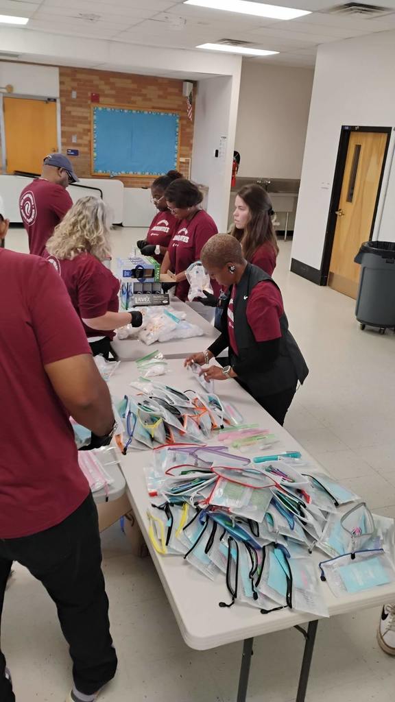 A group of volunteers in maroon shirts are assembling hygiene kits at a table inside a community center. The table is covered with mesh zip pouches, each containing toothbrushes, masks, and other hygiene items. Volunteers are focused on organizing and packing the kits, wearing gloves and working together efficiently.