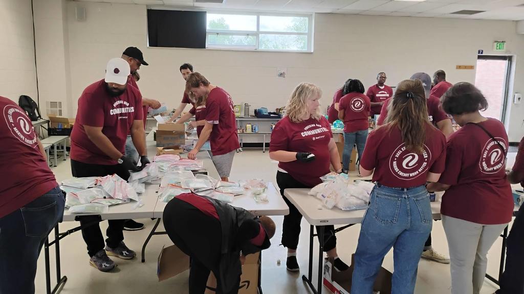 A larger group of volunteers wearing maroon shirts with a circular community service logo work together in a bright room with large windows. They are assembling hygiene kits at several tables, filled with pouches, supplies, and plastic bags. Some people are bending over to pick items from boxes while others are packing on the tables.