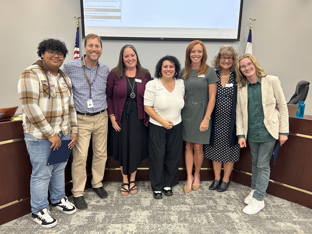 Two students stand at each end of a group photo with the Board of Education; diverse in age, gender, and ethnicity; flags and screen in background.