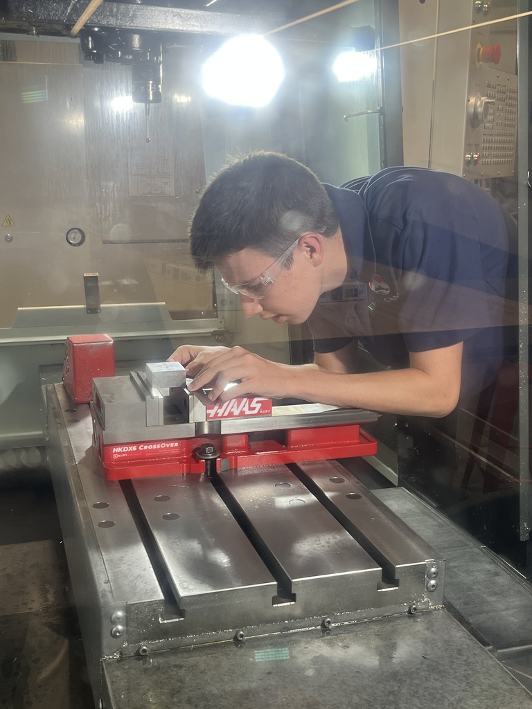 Student in safety glasses and a navy blue "EPIC Campus" shirt operates a CNC machine. The student leans over a red Haas-brand metal vise inside the enclosure, carefully positioning a metal part under bright overhead lights.