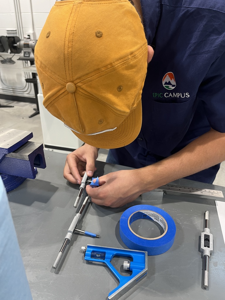 Student wearing a yellow cap and a navy blue "EPIC Campus" shirt uses precision tools at a metal workbench. The student is working with tap wrenches and blue painter's tape. A blue angle square and metal ruler are also on the table. Industrial equipment is visible in the background.