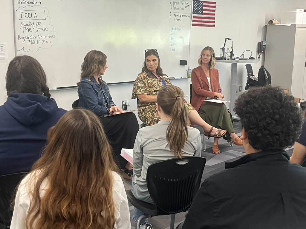 Three women sit at the front of a classroom, leading a discussion with a group of high school students. The women, wearing professional attire, sit facing the students in front of a whiteboard that displays handwritten notes and an American flag. Students are seated in rows, listening attentively.