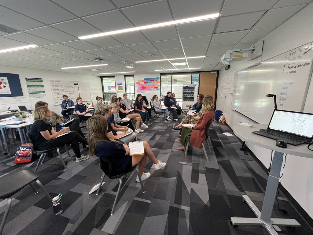 Wide view of a classroom with around twenty students and three women seated at the front leading a panel discussion. Students take notes while facing the panelists. The room has modern furniture, large whiteboards, colorful posters, and a visible laptop displaying notes at the front right.