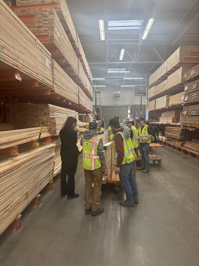 Wide shot of a group of students and adults wearing safety vests in a lumber aisle of a hardware store or warehouse. They are gathered around a flat cart loaded with wooden doors. The space is lined with tall stacks of lumber on metal racks, and a "Restrooms" sign is visible in the background.