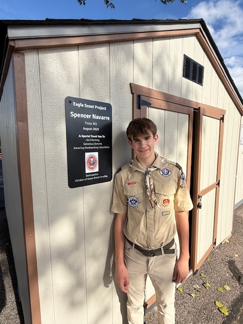 The same scout in uniform stands smiling beside the shed, next to the plaque recognizing his Eagle Scout project.