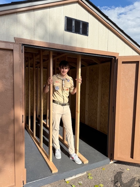 A young scout in uniform stands inside a partially framed section of a newly built shed, holding onto the wooden beams, smiling at the camera.