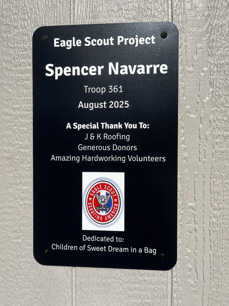 A close-up of a black plaque mounted on the outside of a shed that reads: Eagle Scout Project, Spencer Navarre, Troop 361, August 2025. A Special Thank You To: J & K Roofing, Generous Donors, Amazing Hardworking Volunteers. Dedicated to: Children of Sweet Dream in a Bag. The Eagle Scout emblem is also displayed.
