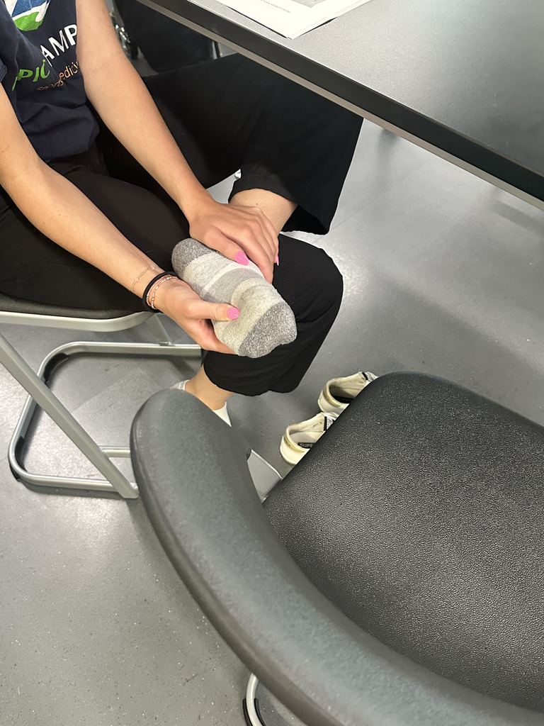 A person seated at a high table in a classroom or lab setting, wearing black pants and a dark navy shirt with a colorful logo partially visible. They are holding their left foot with both hands, which is wearing a gray and white striped sock. Their nails are painted pink. A pair of white sneakers is placed on the floor nearby. The environment includes gray flooring and black chairs with metallic legs.