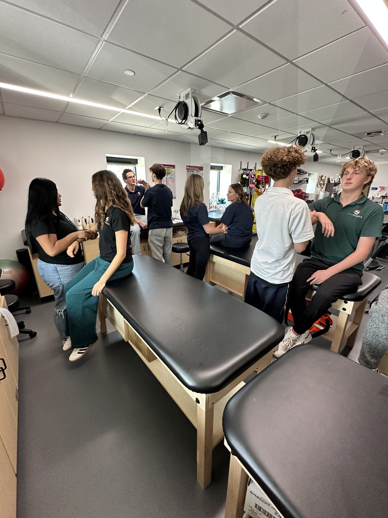 A group of high school students in a medical or sports science classroom, interacting in pairs around several black treatment tables. Some students are standing while others are seated on the tables, appearing to practice physical assessment or therapy techniques on each other's arms or shoulders. The classroom has white walls, a drop ceiling with lights, mounted equipment, anatomical posters, and medical supplies along the back wall. The students are diverse in gender and appearance, and they wear casual or uniform-style clothing.
