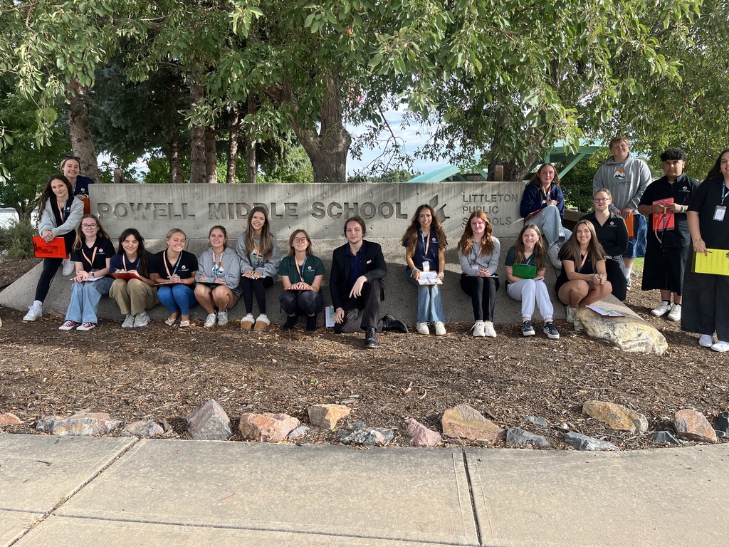 A diverse group of high school students and adults posing in front of a large concrete sign that reads "Powell Middle School – Littleton Public Schools." The group is seated and standing along the sign, smiling and holding colorful clipboards or notebooks. They are dressed casually, some wearing school or program polos and ID lanyards. The background includes tall trees and a partly cloudy sky, giving the scene a welcoming and natural atmosphere.