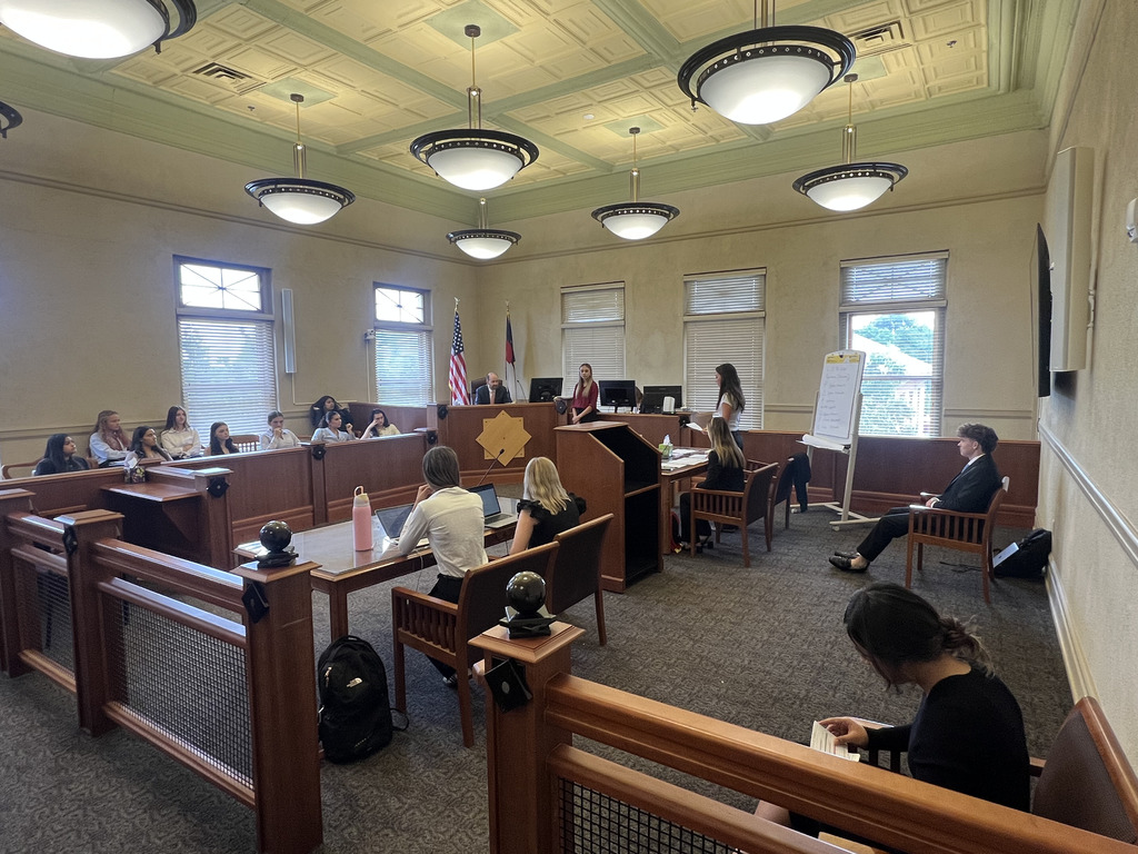 A wide view of a mock trial in progress inside a formal courtroom. Students are seated in various courtroom roles including jury box, judge’s bench, witness stand, and counsel tables. A student stands and presents at the podium, while others take notes. An adult, possibly the judge or facilitator, sits behind the bench near U.S. and Colorado flags. The courtroom features wood furnishings, high ceilings, and hanging circular light fixtures.