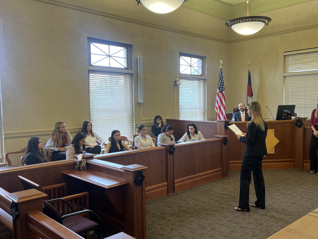 A student in formal attire stands and reads from a paper, presenting to a group of students acting as the jury. The jury members, mostly girls, are seated attentively in the jury box. Behind the student is a judge’s bench with an adult smiling and watching the proceedings. U.S. and Colorado flags are visible beside the bench. The setting is a traditional courtroom with wood paneling and large windows.
