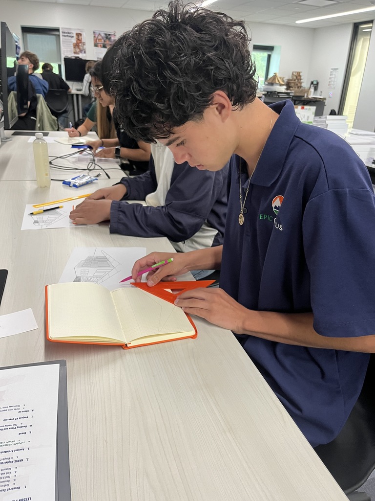 A student with curly dark hair wearing a navy "EPIC Campus" polo shirt sketches with a pencil and orange triangle ruler in a graph notebook. He is seated in a classroom at a long desk beside other students also working on architectural sketches. A bottle, eraser, and other supplies are on the desk.