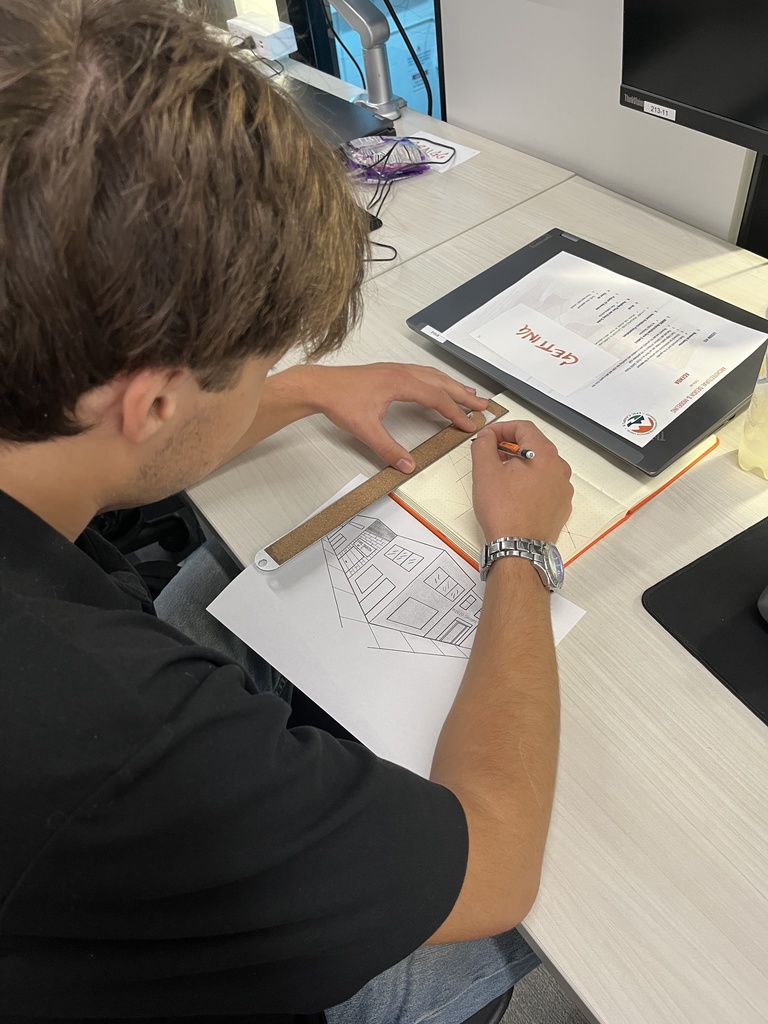 A student with short brown hair wearing a black shirt uses a ruler and pencil to draw a perspective sketch of a building on graph paper. A printed lesson plan and tablet with "LPS" branding are on the desk. The student is seated at a modern workspace with a monitor and desk lamp visible.