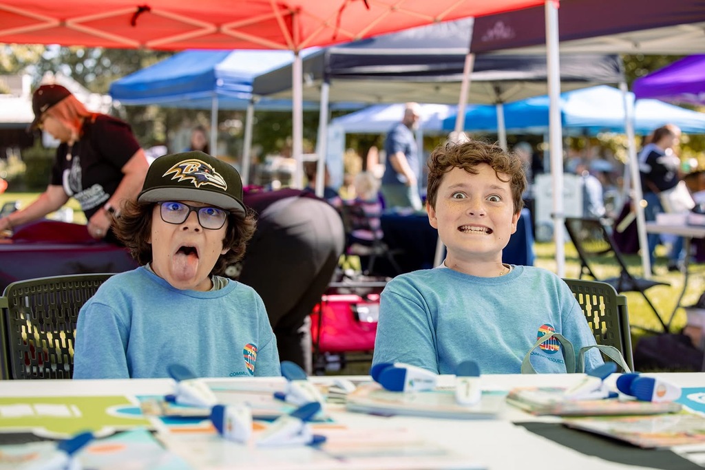 wo children wearing light blue shirts sit at a booth table under colorful tents at the community resource expo 2024. One child is making a silly face with their tongue out and wearing a black baseball cap, while the other is smiling wide with a playful expression. Flyers and small giveaway items are spread out on the table in front of them.