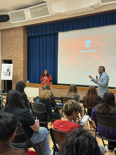 Dr. José Enriquez, founder of Latinos in Action, stands on stage speaking to students. A student in a red shirt joins him in addressing the audience seated in rows of chairs.