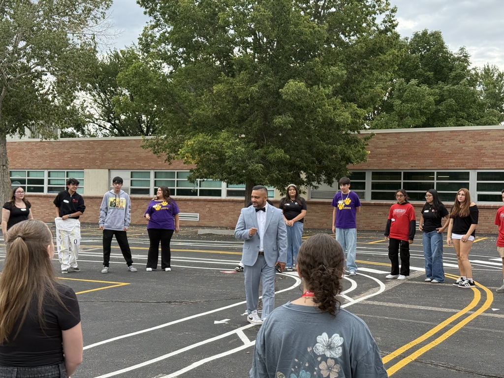 Dr. José Enriquez, founder of Latinos in Action, speaks outdoors to LIA students gathered in a large circle on the playground, with trees and a brick school building in the background.