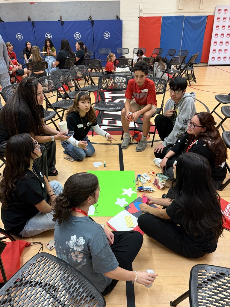 Latinos in Action students and an adult sit in a circle on the gym floor, working on a bright green poster with paper cutouts, markers, and craft supplies.