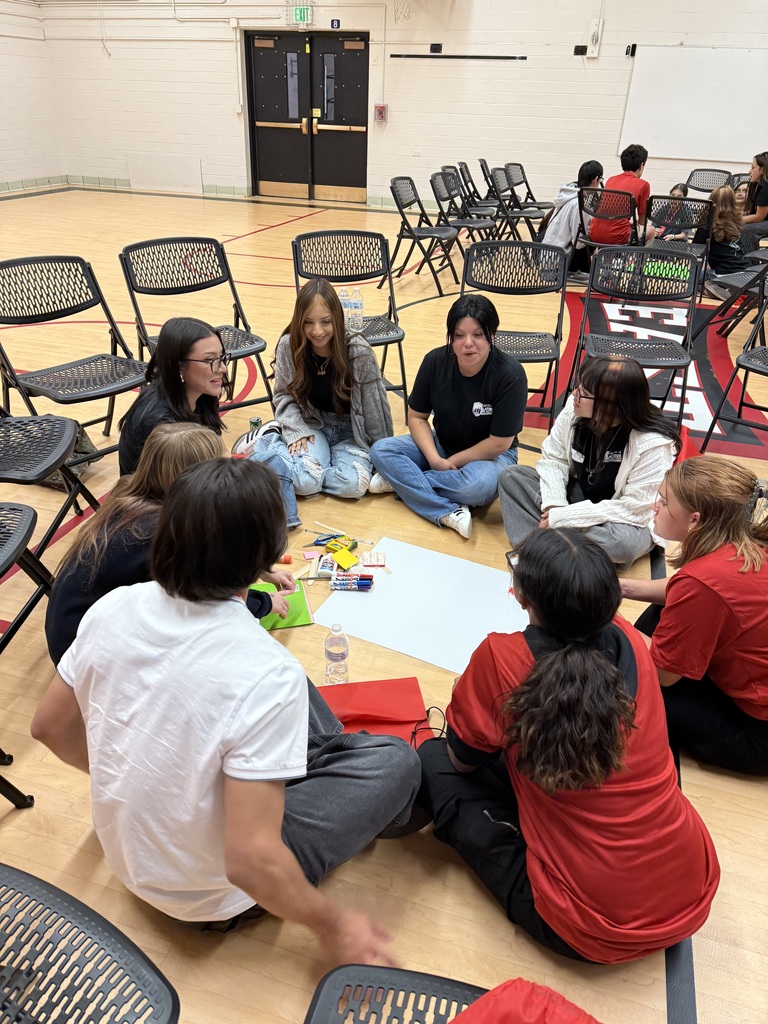 Latinos in Action students and an adult sit in a circle on the gym floor, working on a bright green poster with paper cutouts, markers, and craft supplies.