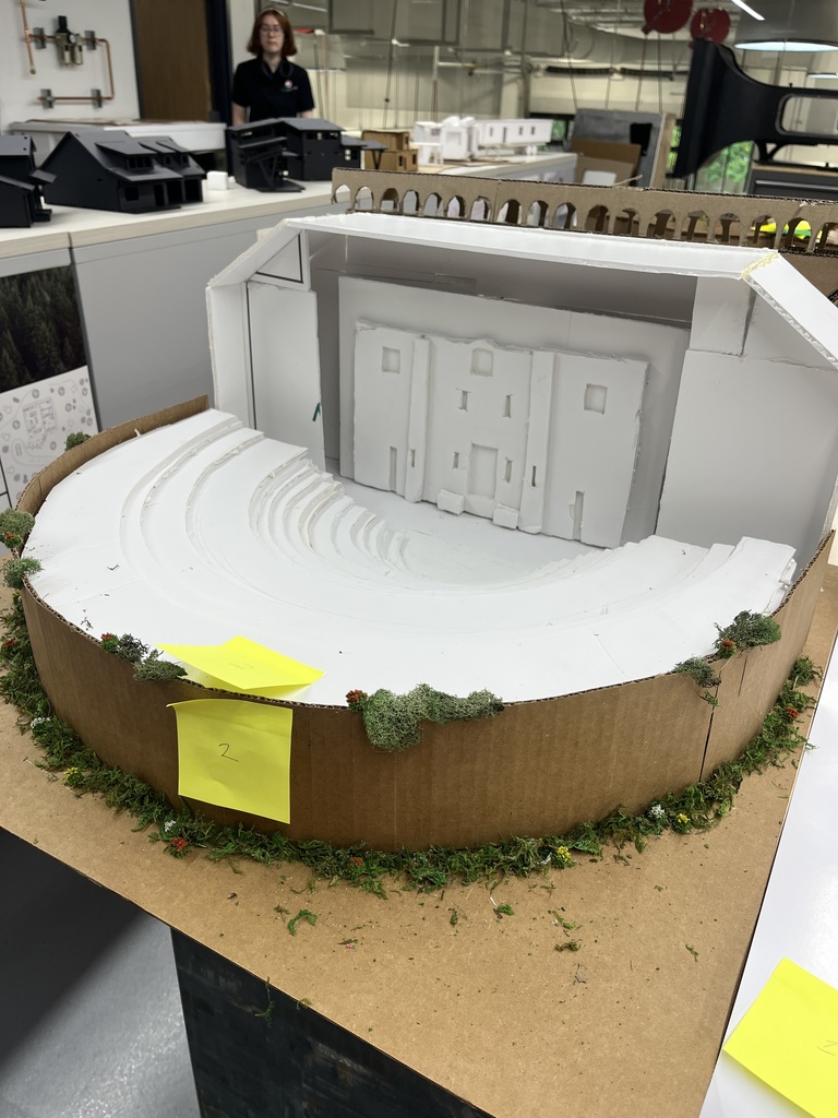 A handmade scale model of an ancient Roman-style amphitheater, built from white foam board and cardboard. The semi-circular seating area features tiered rows leading to a central stage with a rectangular backdrop that resembles a building façade. The model includes greenery made from artificial moss and flowers around the outer edge. A yellow sticky note labeled “2” is attached to the front. In the background, a woman stands near other architectural models on display in a classroom or workshop setting.