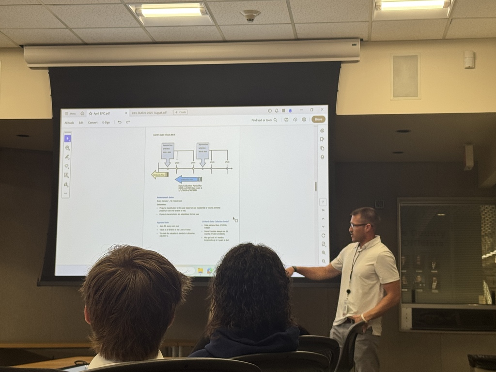 A man in a white polo shirt presents in front of a projected screen showing a PDF document with a flowchart and text. He gestures toward the screen while standing beside seated attendees, including two individuals visible from behind. The setting is a modern conference or classroom space with a drop ceiling and recessed lighting. The visible document on the screen appears to outline dates and deadlines, likely for an academic or administrative process.
