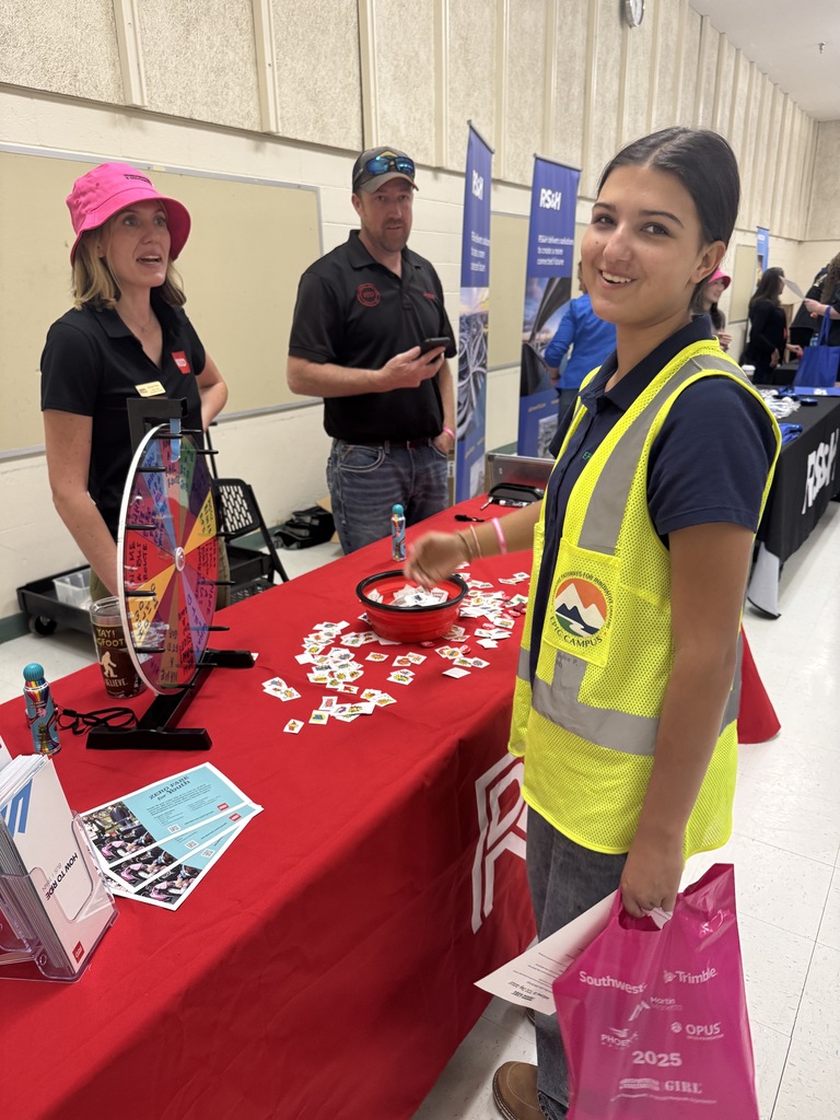 A student smiles at the camera while standing at a booth with a red tablecloth covered in small paper tokens. She wears a yellow safety vest with a logo reading “Construction for Kids” and carries a bright pink event bag. Two adults in black shirts, one wearing a bright pink hat, stand behind the table, which features a colorful prize wheel and brochures. Several informational banners are displayed behind them. The setting is a school or community career fair.