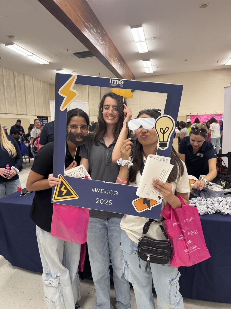 Three young women pose inside a large cardboard photo frame labeled “ime” and “#IMExTCG 2025” at a school or community event. They wear playful props such as paper sunglasses, a beard, and a light bulb cutout. All three are smiling, and two are holding papers and bright pink event bags. A busy room with other attendees and booth tables is visible in the background.