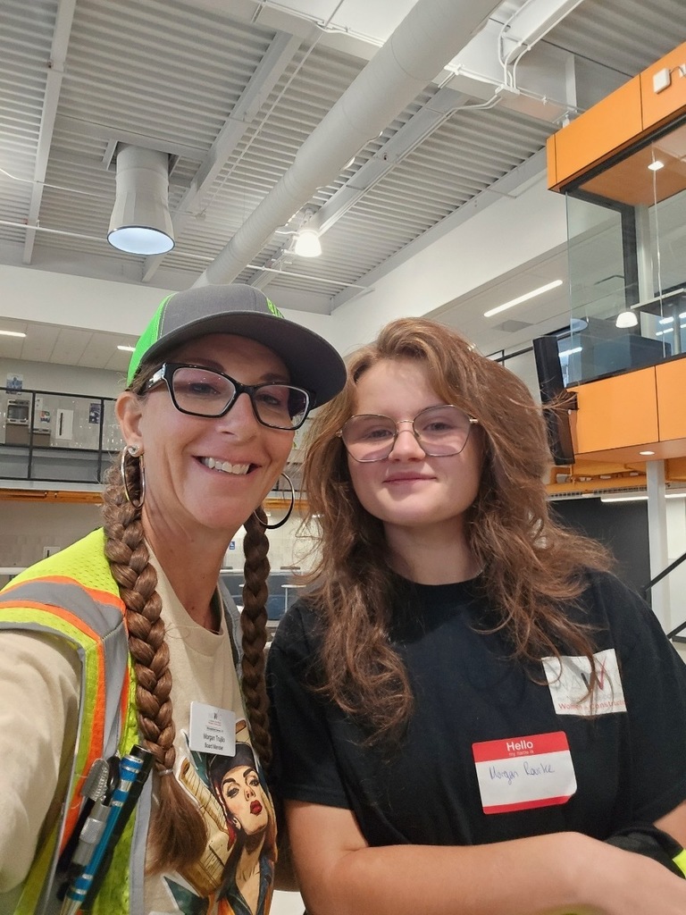 Two individuals pose for a selfie in a bright, modern indoor space with exposed ceilings and large windows. The adult on the left wears glasses, a green and gray cap, and a construction safety vest over a t-shirt with a Rosie the Riveter-style print. Her name tag reads “Megan Rado, Board Member.” The student on the right wears glasses and a black t-shirt with a name tag that says “Morgan Baske.” Both are smiling.