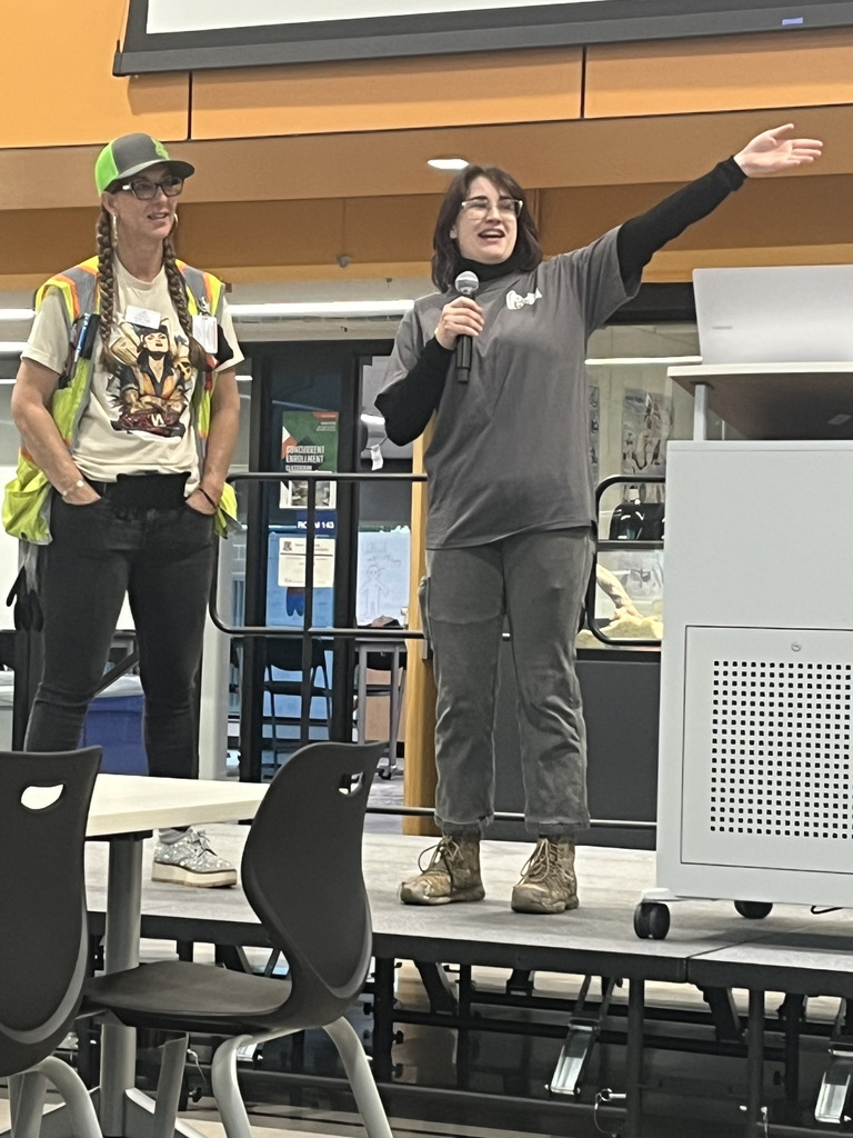Two women stand on a raised platform in a classroom or cafeteria-like setting. One wears a safety vest and graphic t-shirt, the other holds a microphone and speaks while smiling. They both wear name tags, and behind them is a projector screen and a bright orange structure. Empty black chairs and tables are in the foreground.