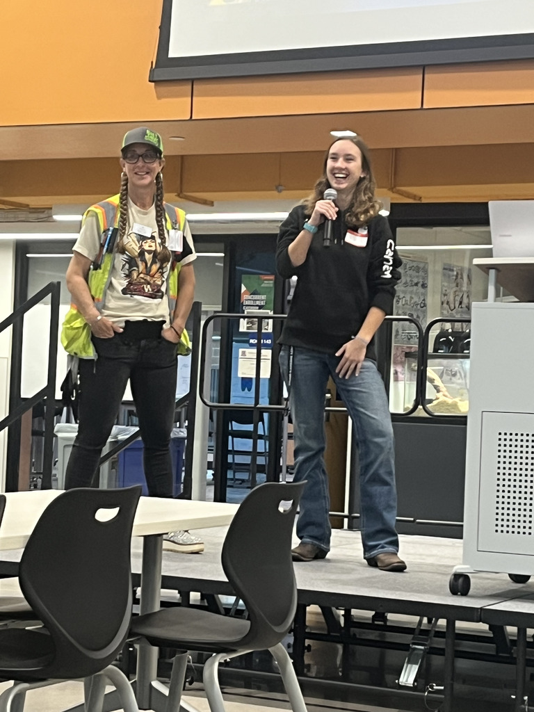 Two women stand on a platform in a large room. One, in a safety vest and colorful t-shirt, stands with hands in pockets. The other woman, wearing a gray t-shirt and camouflage-style boots, holds a microphone and gestures outward while speaking. Classroom furniture and a projector setup are visible in the foreground.