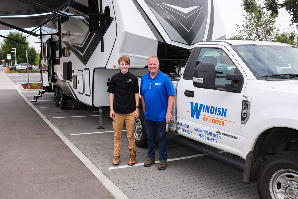 Two smiling people stand in front of a large white RV and a white Windish RV Center truck. One is a teenage boy wearing a black button-up shirt with a logo and tan pants; the other is an adult man wearing a blue Windish RV Center polo and dark jeans. The truck has the Windish RV Center logo and contact information for Longmont, Lakewood/Denver. The RV is parked with extended awnings in a paved parking lot bordered by trees and street signs in the background.